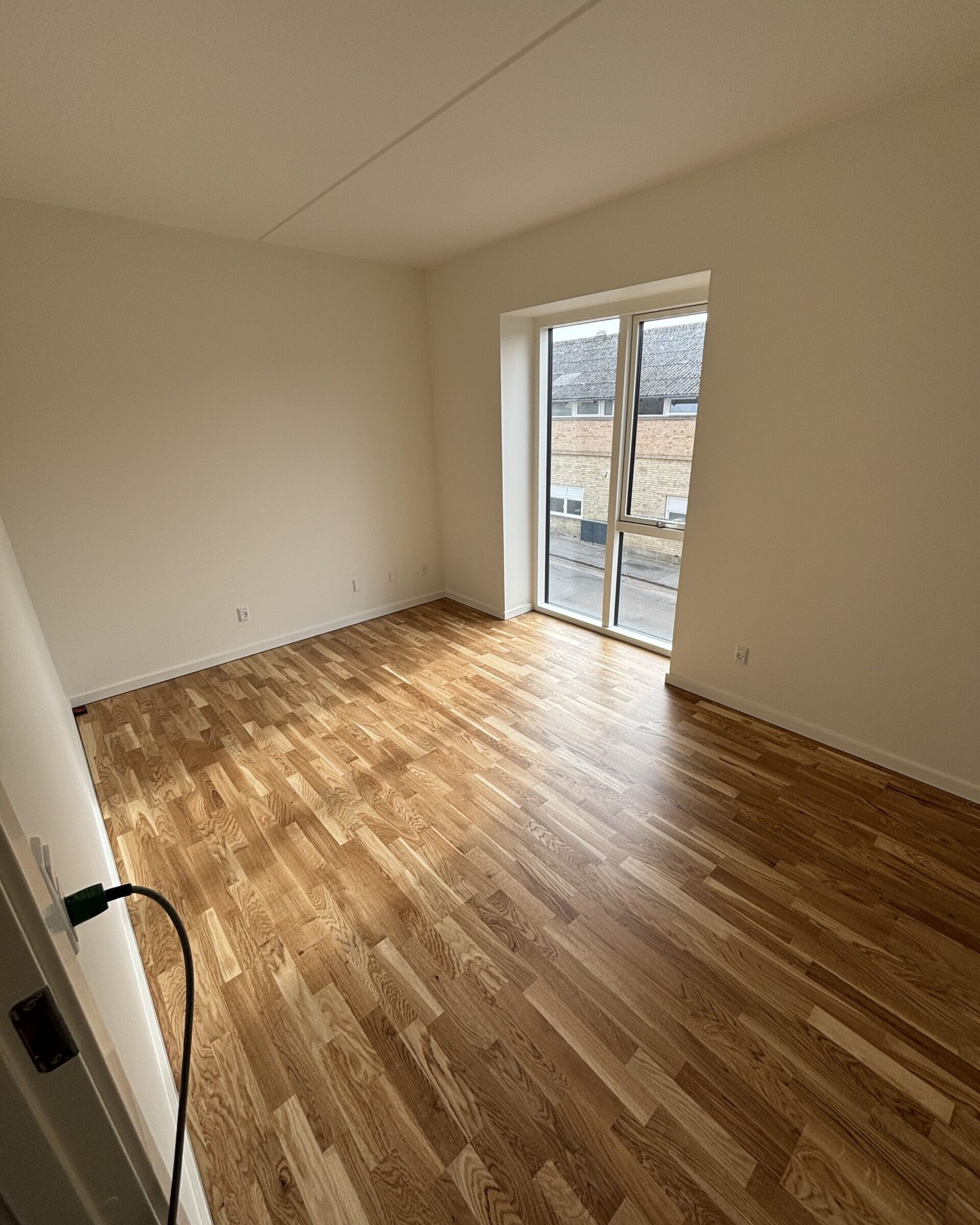 Empty room with light wood flooring and a large window letting in natural light; white walls.