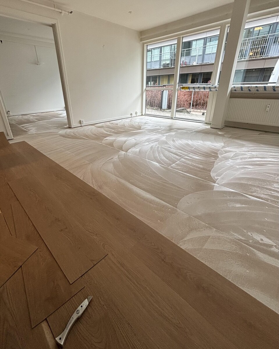 Room under renovation with new light wood laminate planks being installed; protective white underlayment covers most of the floor and a few planks lie stacked on the left. Large windows and a balcony overlook a neighboring building.