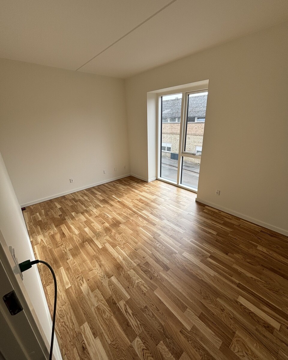 Bright empty room with light wood floor and a tall window letting in natural light. The window overlooks a brick building outside.