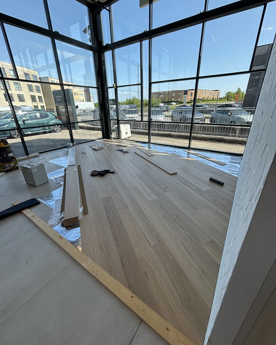 Interior of a glass-walled room under construction with new wooden flooring being installed and tools on the floor.