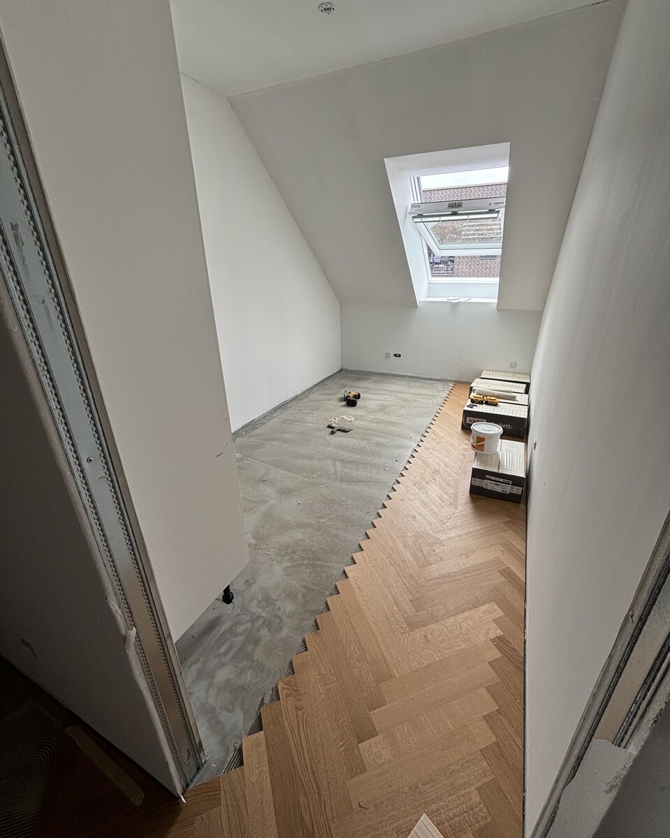 Under-construction attic room with sloped ceiling, a large skylight window, and new wooden parquet flooring being installed; tools and boxes nearby.