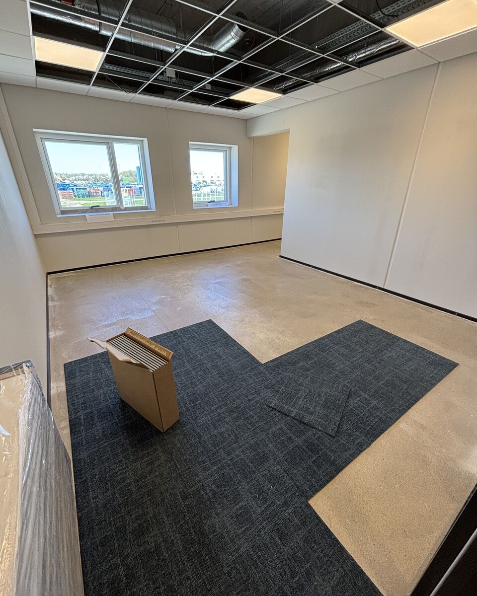 Construction‑phase office space with exposed ceiling, two front windows, and scattered dark carpet tiles; a cardboard box rests on the floor.