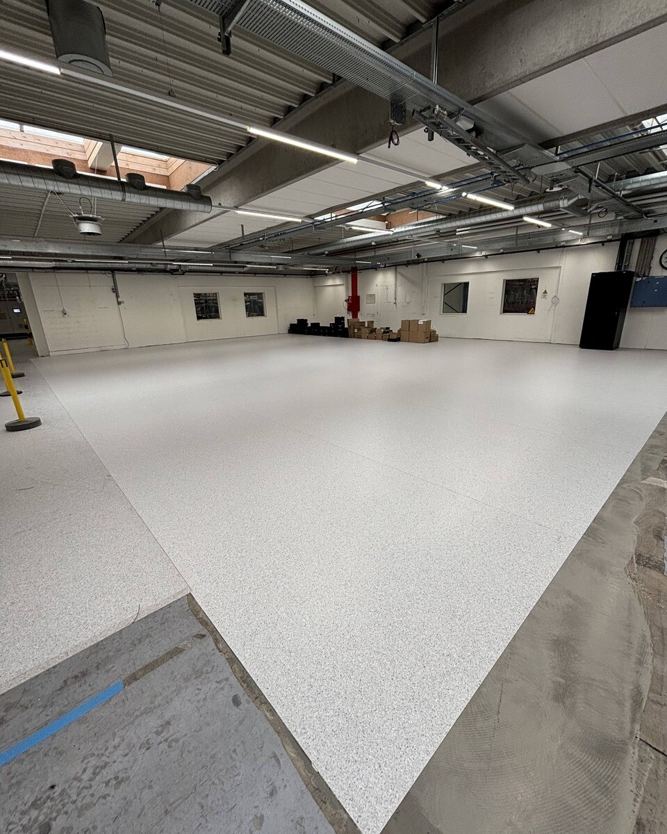 Bright empty industrial workspace with a speckled epoxy floor, exposed ceiling ducts, and boxes along the far wall.
